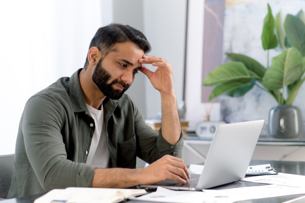 Upset arabian or indian man, tired, overworked working in a laptop, looks sadly at the screen, is stressed because of a work not done on time, an unsuccessful project, a startup, needs a vacation