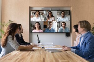 Millennial business team meeting in boardroom, talking on video conference call to happy multiethnic remote distant freelance employees, turning looks to head shots set on big screen