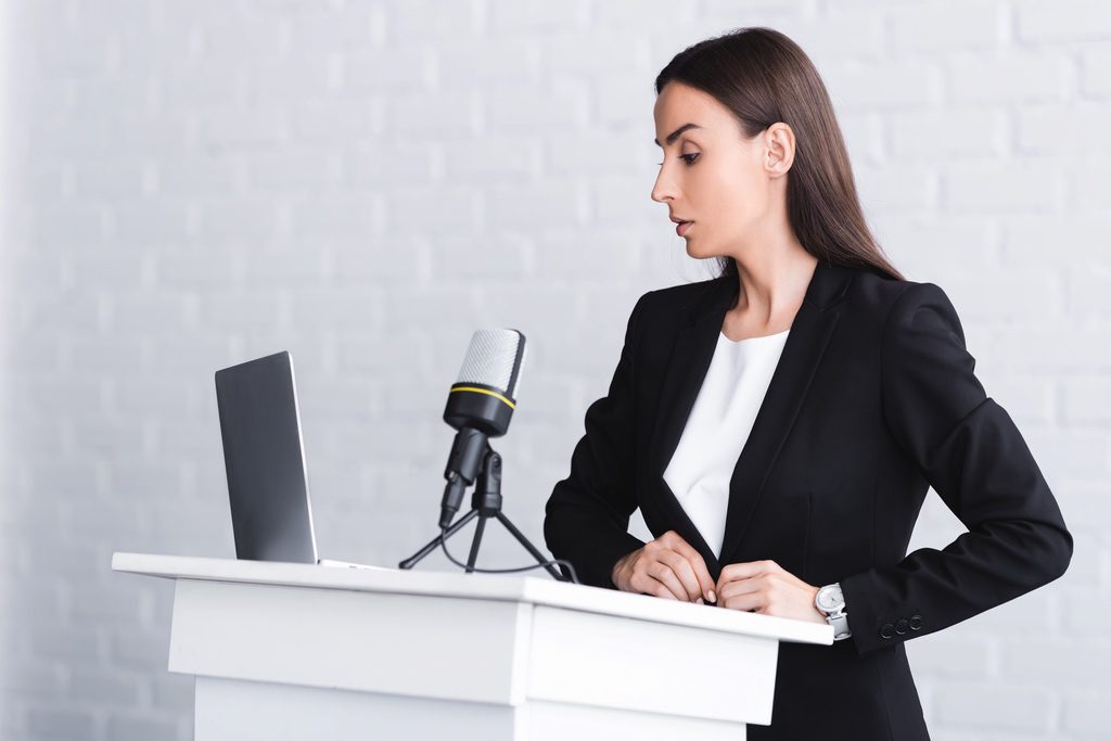 Woman speaking into microphone at podium while looking at laptop screen.