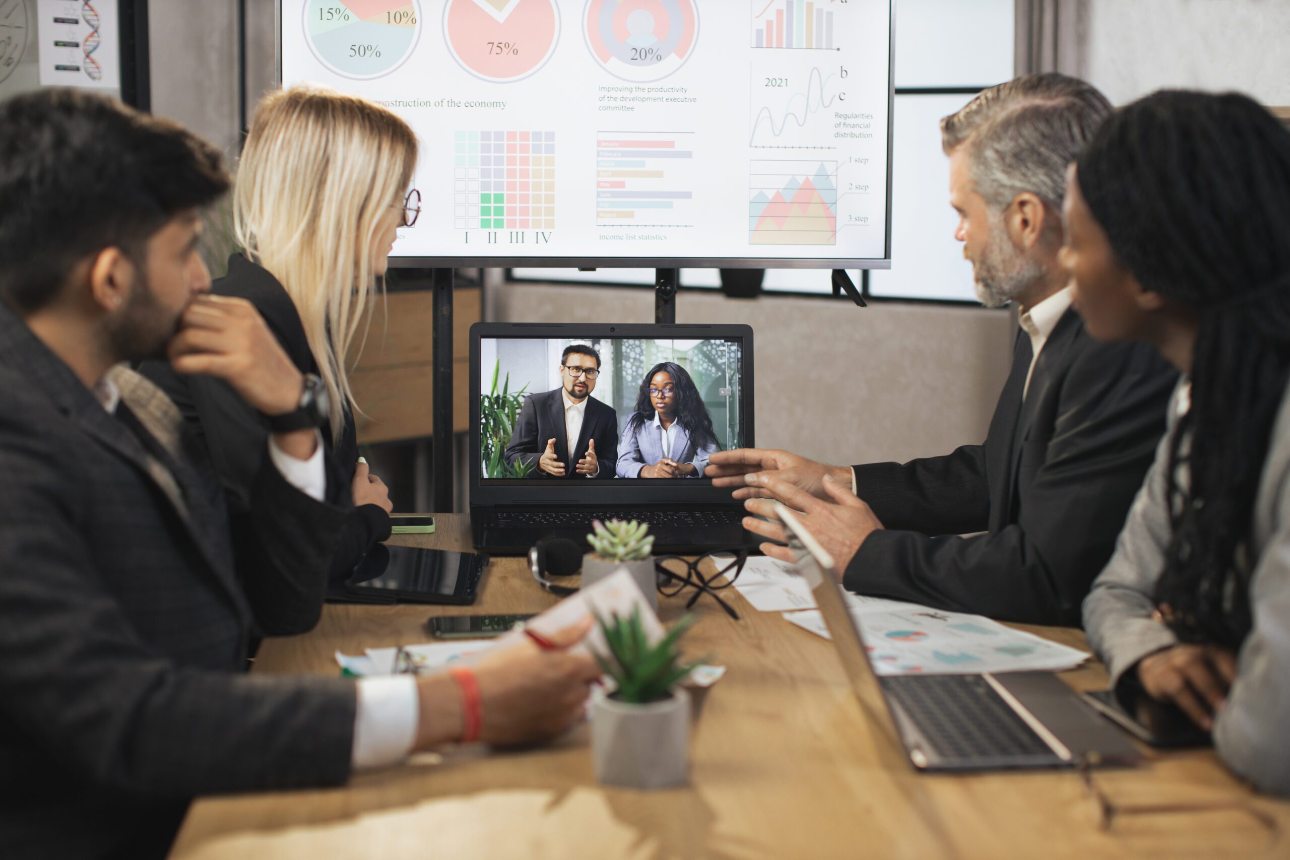 Business professionals in a hybrid meeting, with four people in a board room collaborating with two remote colleagues. 